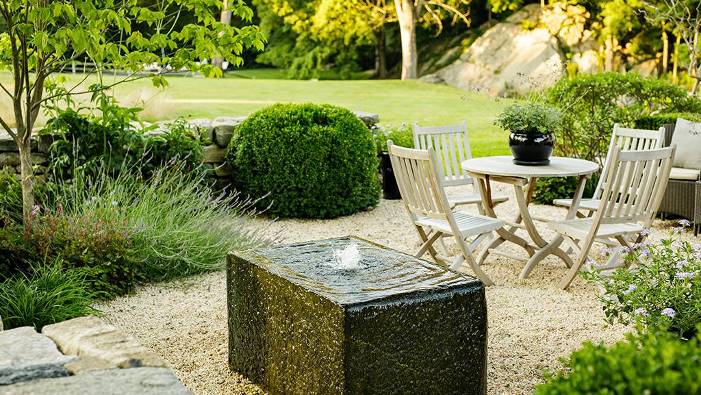 bubbling rectangular water feature in an ourdoor dining room with a table and four chairs in background and green landscape surrounding the space