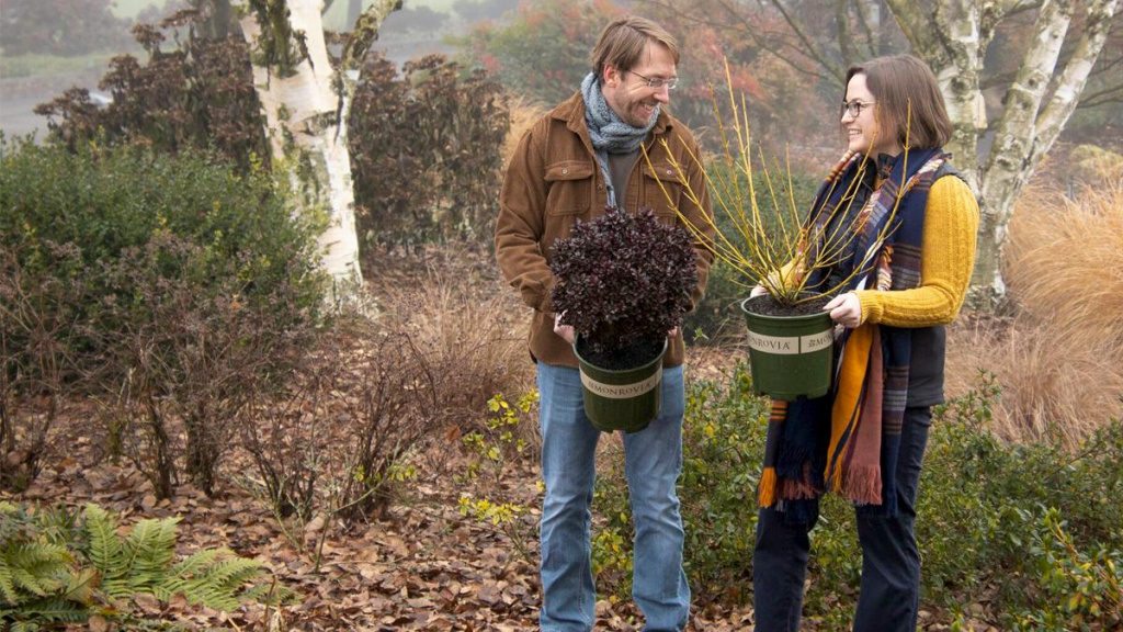 A man and women gardener in a backyard garden each carrying a Monrovia potted plant.