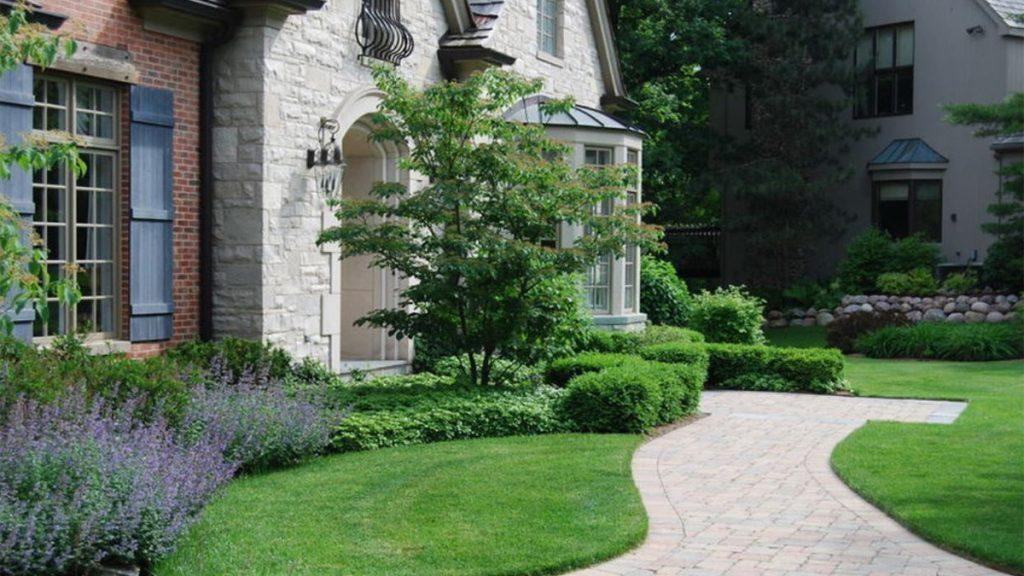Path leading to the front of a house with purple flowers and green grass, bushes, and trees. 